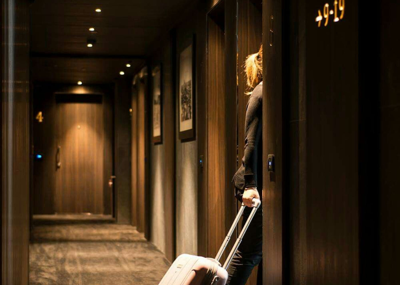 Hotel guest entering a modern luxury hotel room with a rolling suitcase, warm ambient corridor lighting and room numbers illuminated on the wall.