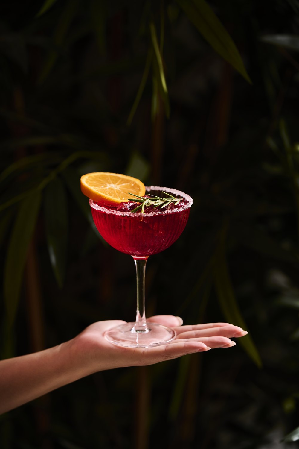 A hand holding a deep red O.W. cocktail flat on an open palm against dark foliage, photographed by JUDE&trade;.
